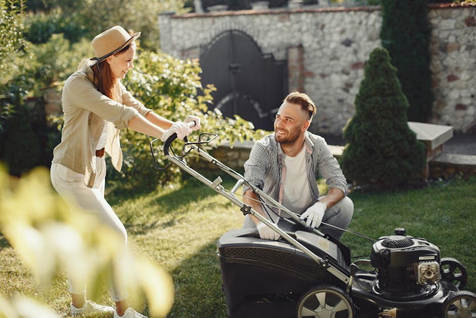A person wearing shorts and sturdy gardening boots operates a petrol-powered lawn mower on a well-maintained grassy lawn during late afternoon sunlight. The grass appears lush and evenly cut, with vis